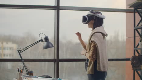 Female-engineer-in-a-virtual-reality-helmet-near-the-large-window-with-his-hands-imitates-the-work-of-the-interface.-Designing-the-future-the-concept-of-virtual-architecture-and-design-interface-graphic-applications.